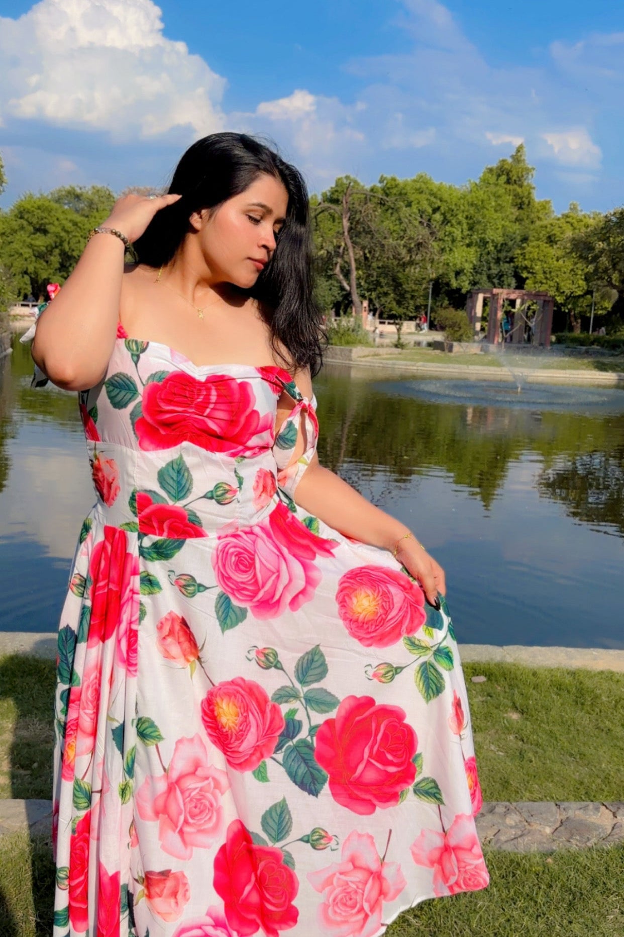 Woman in a floral dress sitting by a pond with trees and blue sky in the background