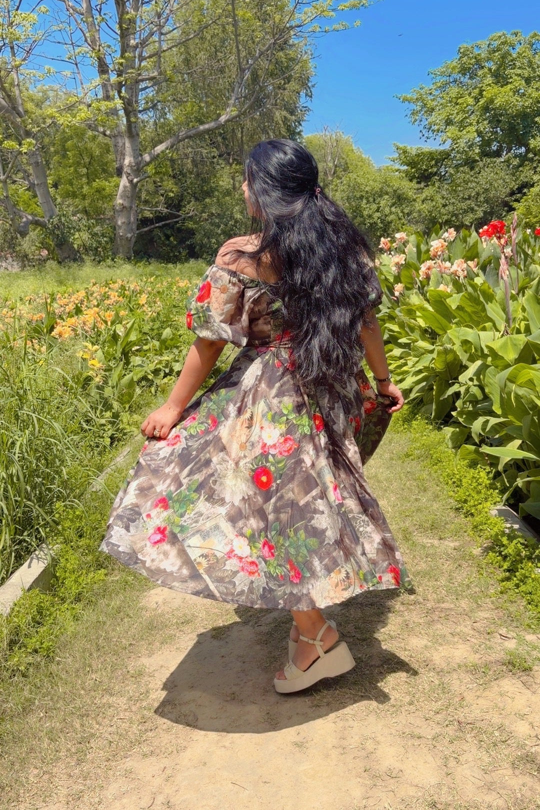 Woman in a floral dress walking through a garden with flowers and trees.